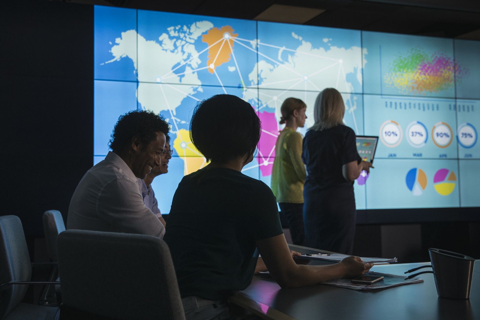 A small multi-ethnic group discusses business decisions in a boardroom by using data visualization techniques to interpret data